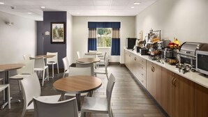 A modern dining area with round tables and white chairs, a kitchen counter with appliances, and a window with blue curtains. - Microtel Inn & Suites By Wyndham Lynchburg (Lynchburg)