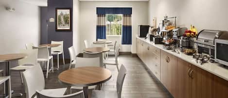 Un área de comedor moderna con mesas redondas y sillas blancas, una encimera de cocina con electrodomésticos, y una ventana con cortinas azules.