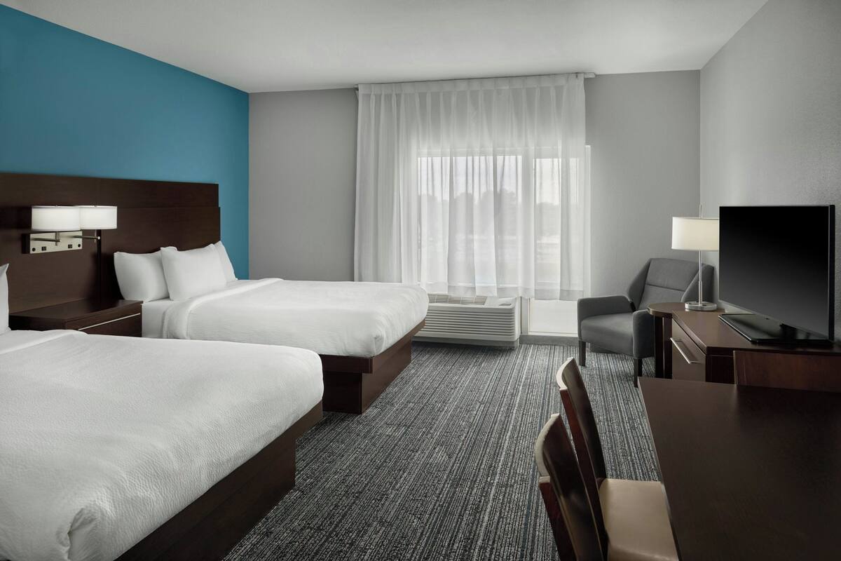 A hotel room with two beds, a desk, a TV, and a large window with curtains.