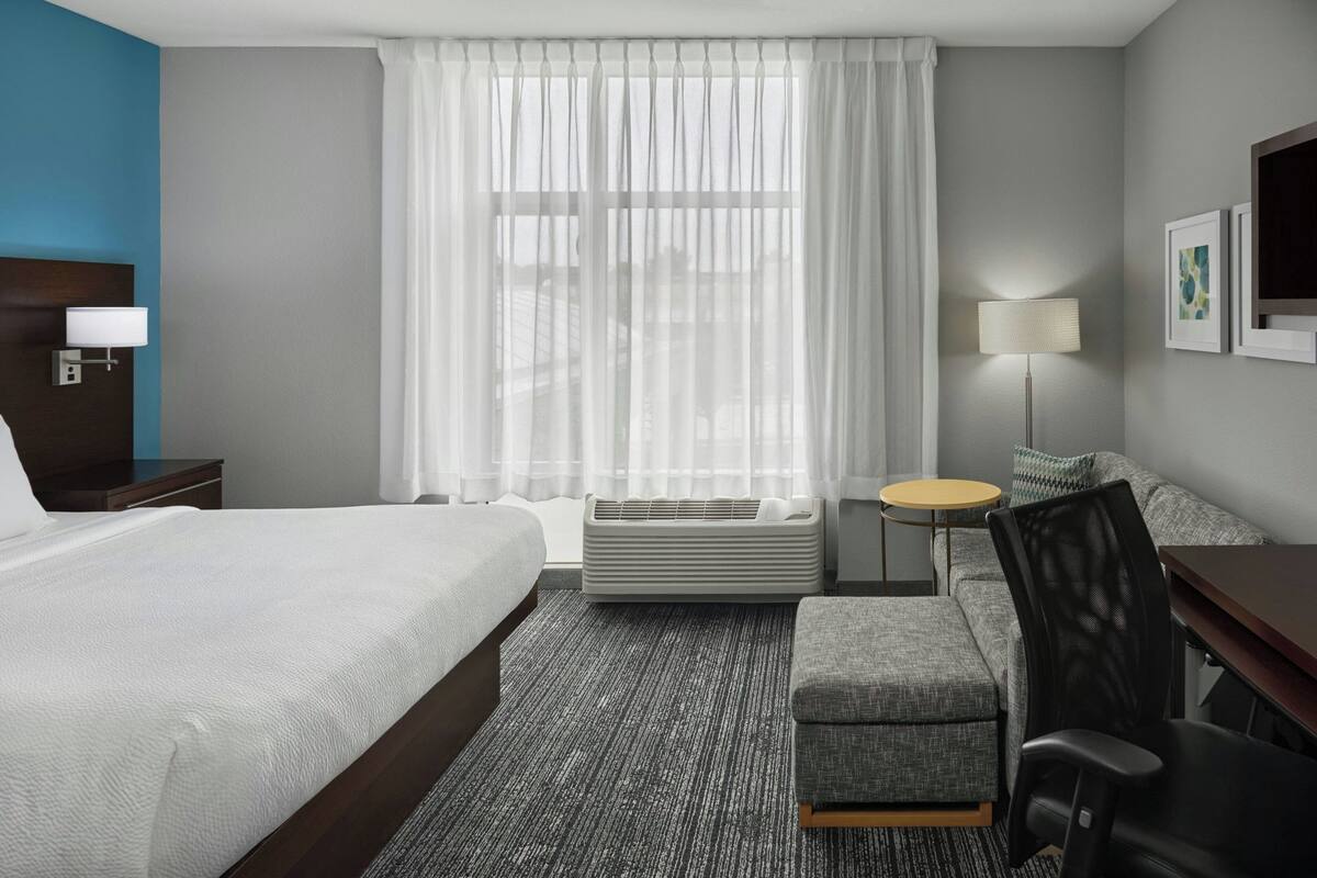 A hotel room with a bed, a desk, a chair, a round table, a lamp, and a large window with sheer curtains.