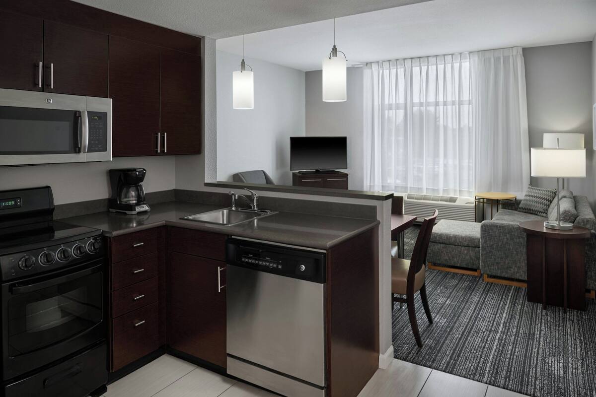 A compact kitchen with a microwave, stove, and dishwasher, a seating area with a sofa and armchair, and a large window with curtains.