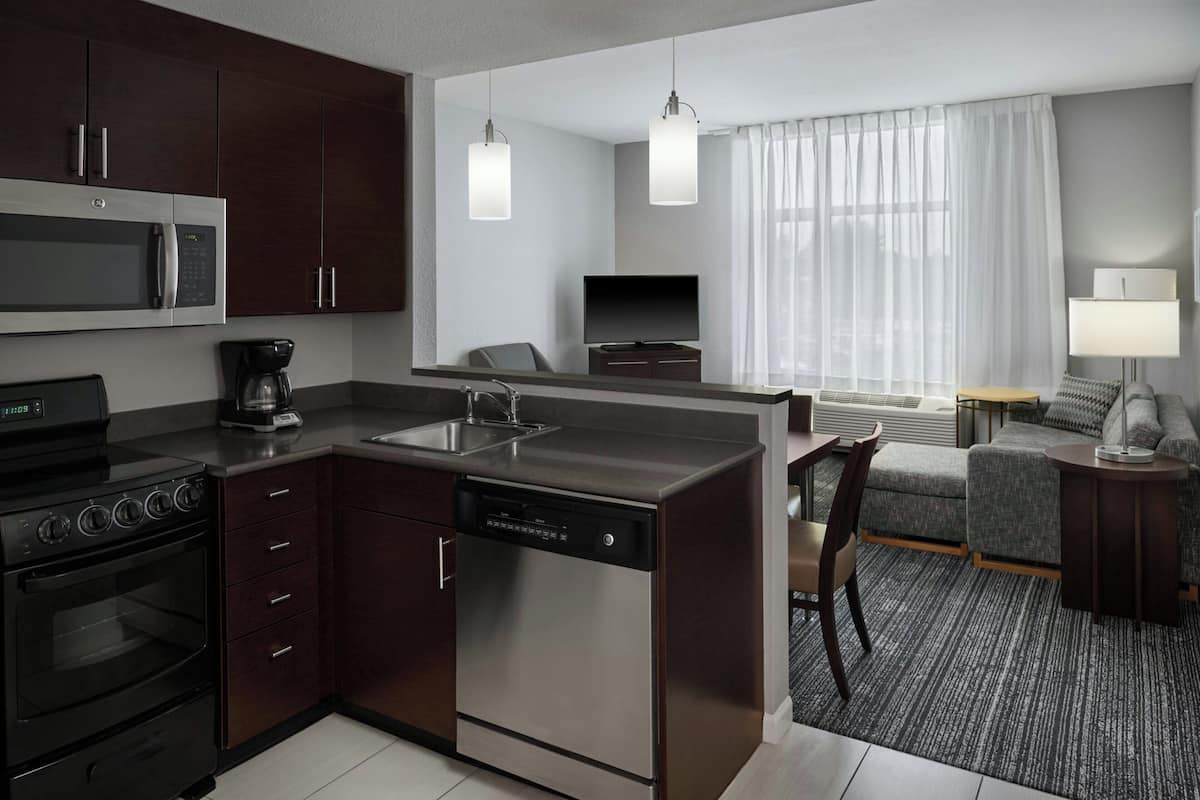 Cocina compacta con microondas, estufa y lavavajillas, zona de estar con sofá y sillón, y un amplio ventanal con cortinas.