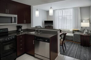 A compact kitchen with a microwave, stove, and dishwasher, a seating area with a sofa and armchair, and a large window with curtains. - TownePlace Suites Springfield (Springfield)