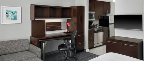 A hotel room with a kitchenette, a desk, a chair, a bed, a sofa, and a flat-screen TV.