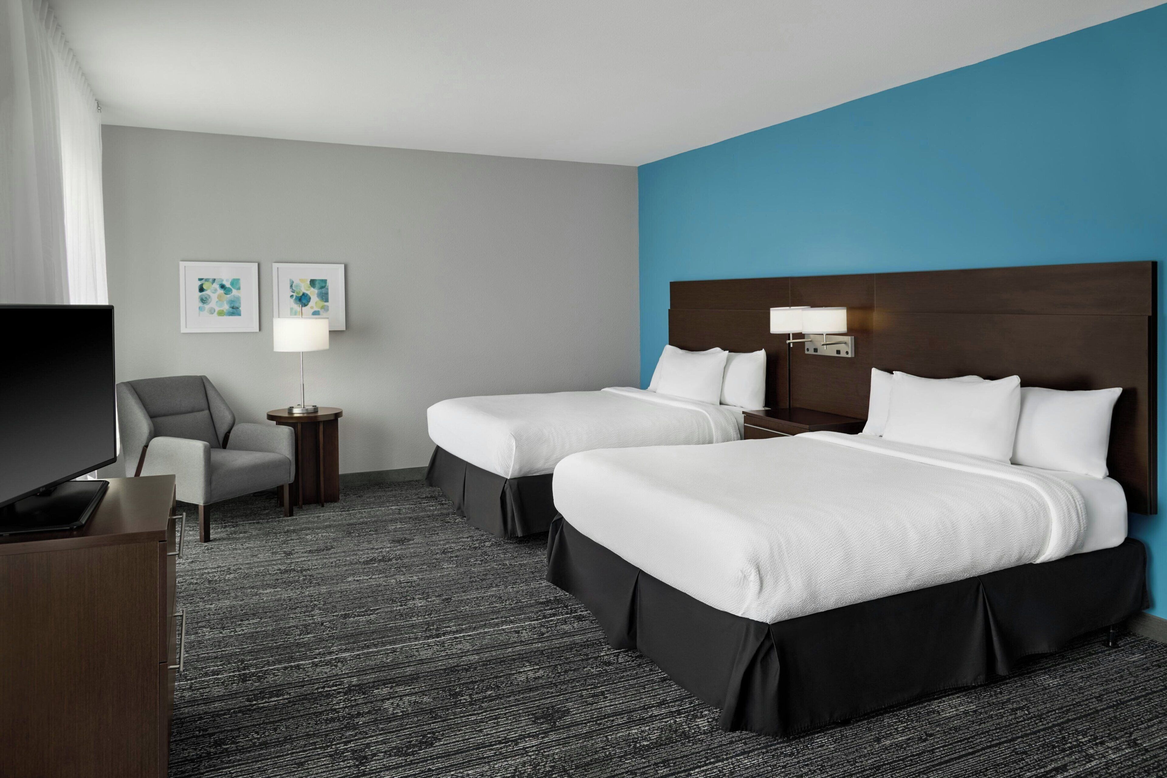 Habitación de hotel con dos camas, televisión, silla, lámpara y cuadros en la pared.