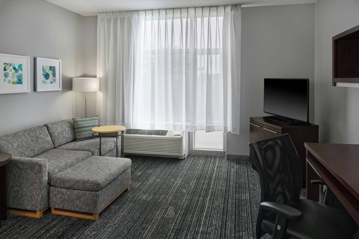 A hotel room with a sectional sofa, a small round table, a TV, and a desk with a chair.