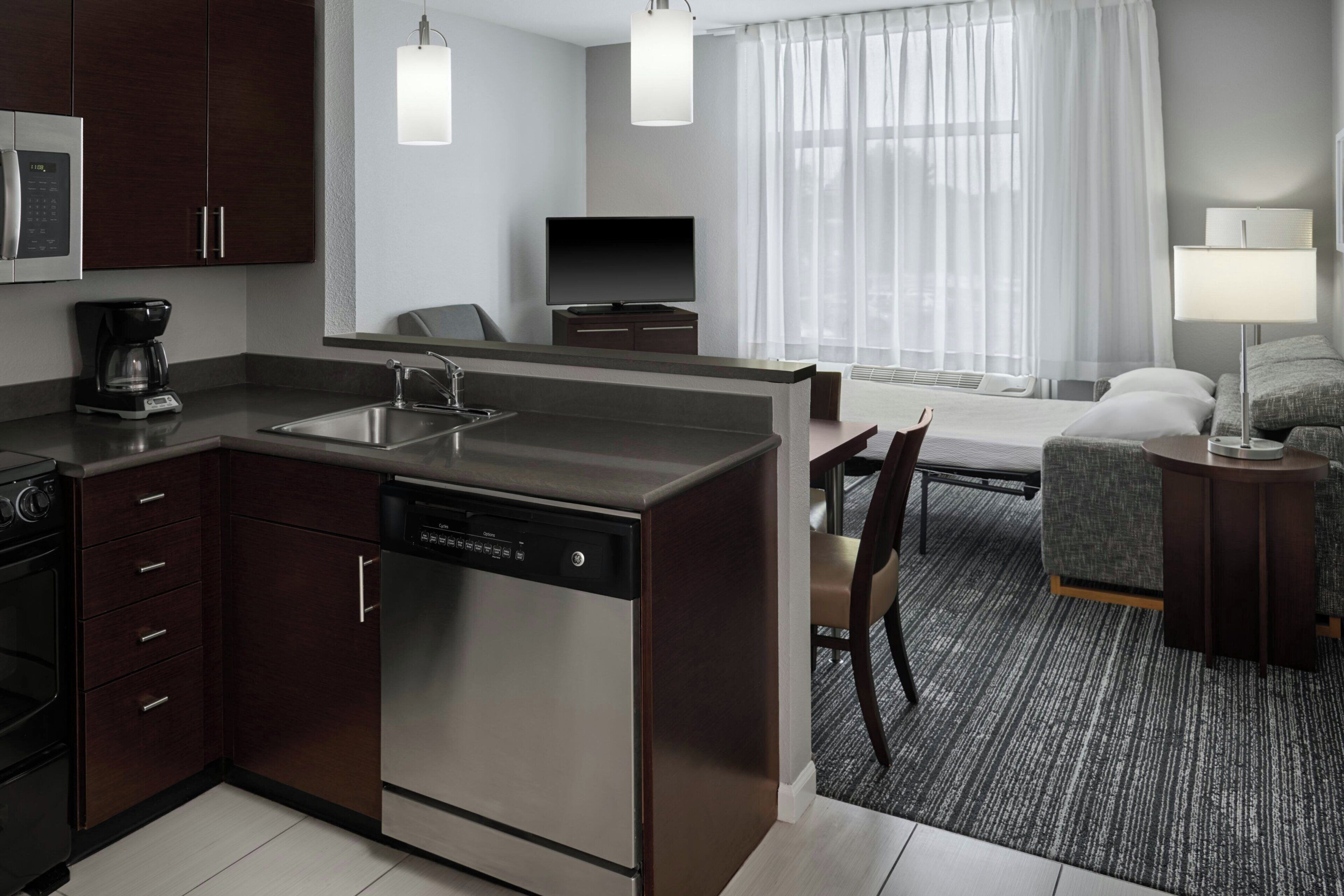 A modern hotel room with a kitchen area, a dining table, a sofa, and a TV.