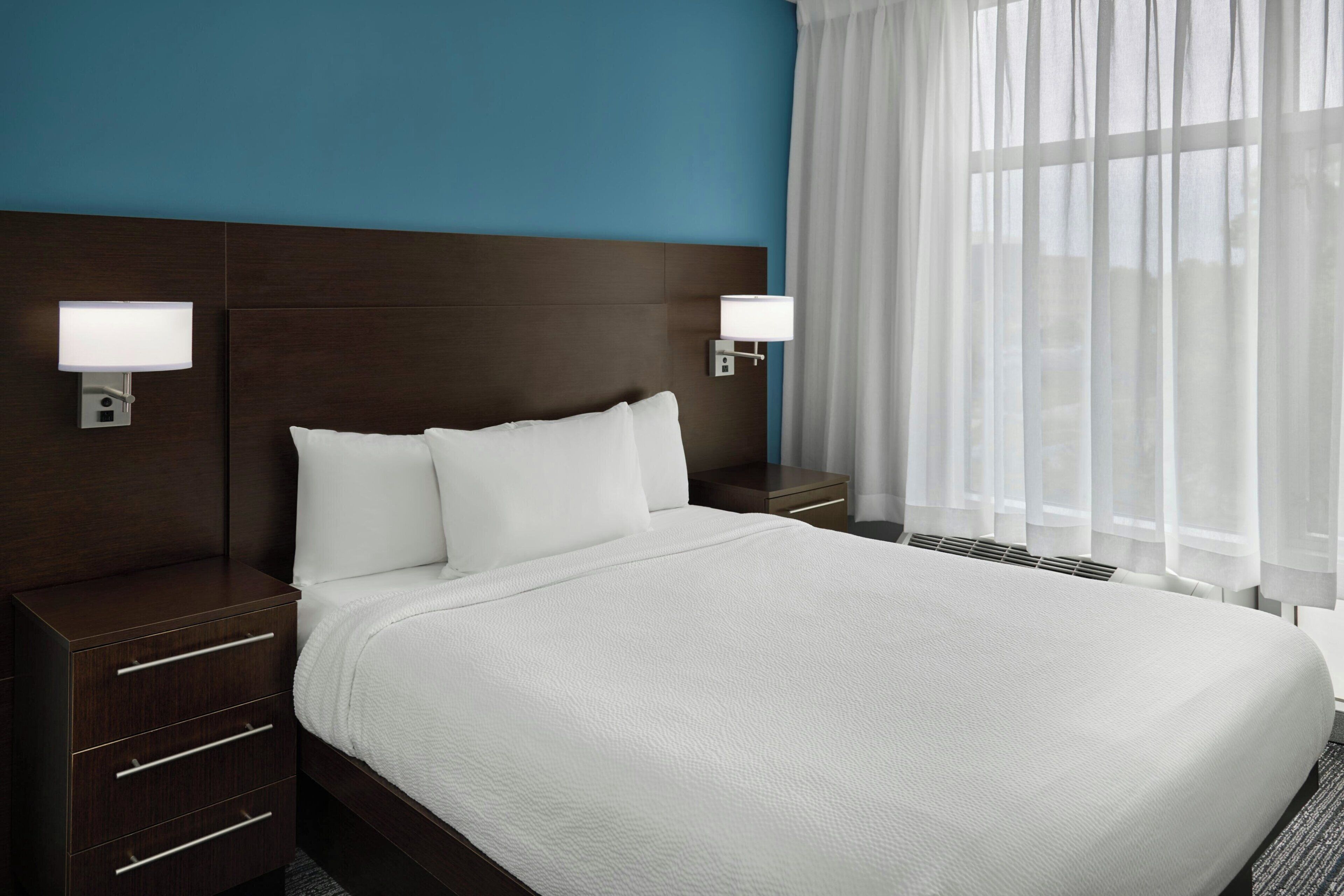 Une chambre d’hôtel avec un grand lit, deux tables de chevet, une petite table et une fenêtre avec des rideaux légers.