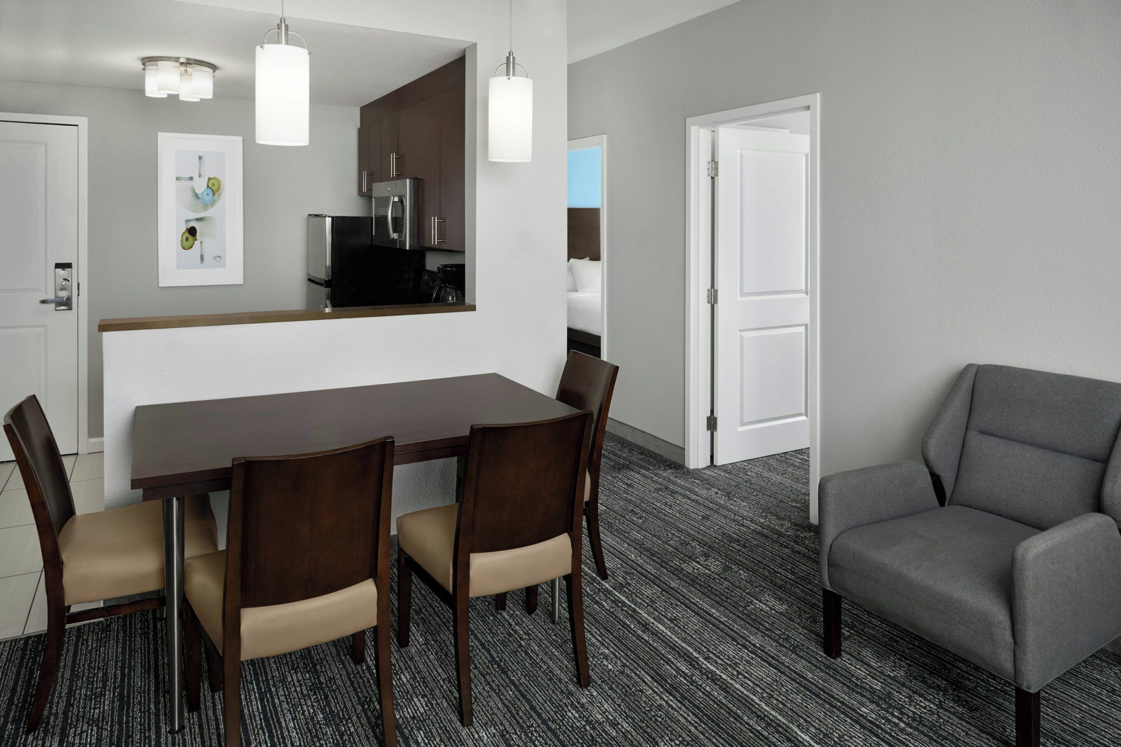 A hotel room with a dining table, four chairs, a gray armchair, a kitchen area, and a white door.