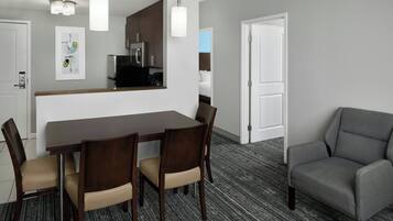 A hotel room with a dining table, four chairs, a gray armchair, a kitchen area, and a white door.
