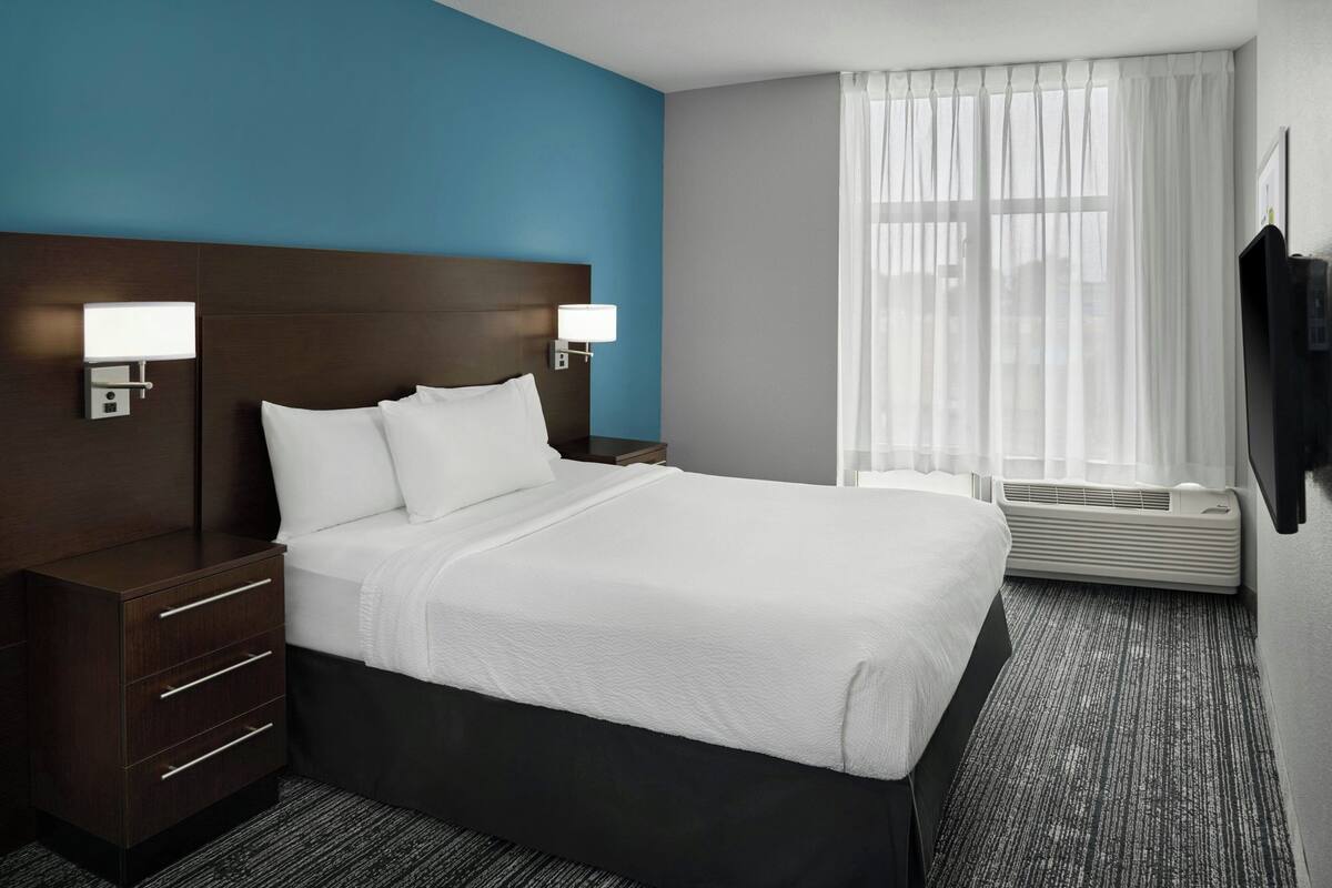 A hotel room with a bed, bedside tables, lamps, a window with curtains, and an air conditioning unit.