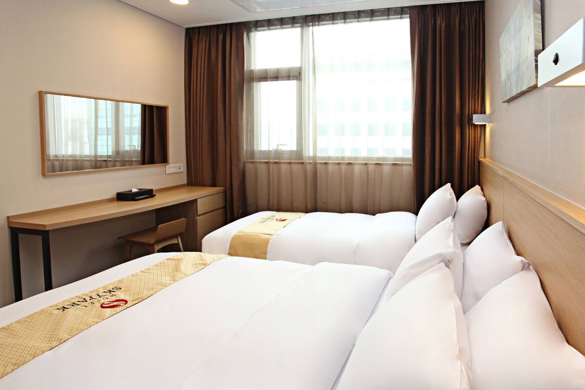 Une chambre d’hôtel avec deux lits, un bureau, une chaise, une grande fenêtre avec des rideaux et un luminaire fixé au mur.