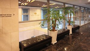 Un vestíbulo de hotel con mostrador de recepción, un cartel que indica la sala de reuniones y la lavandería, y dos plantas en macetas con un banco delante.