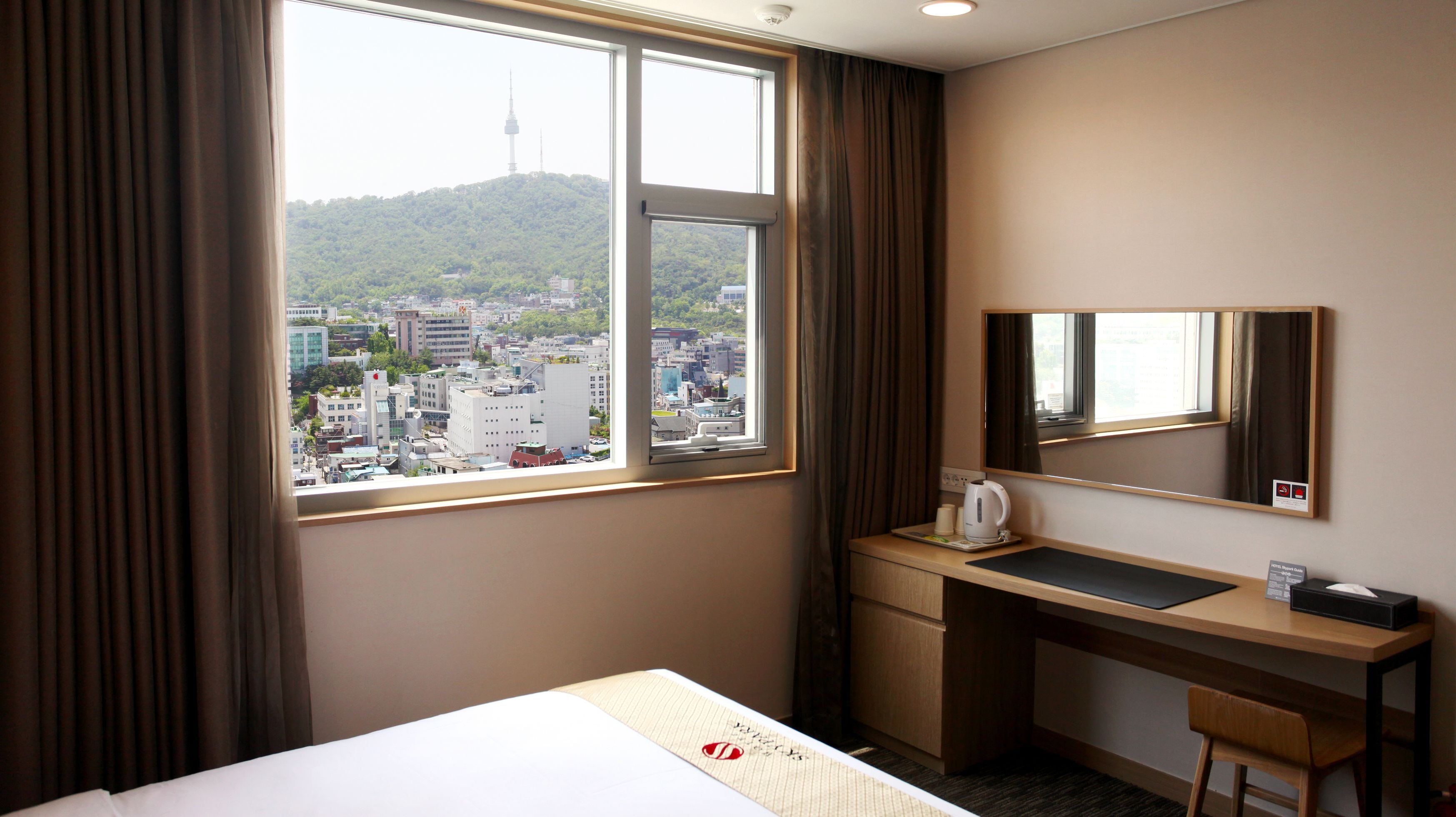 A hotel room with a large window, a desk, a chair, a bed, and a mirror.