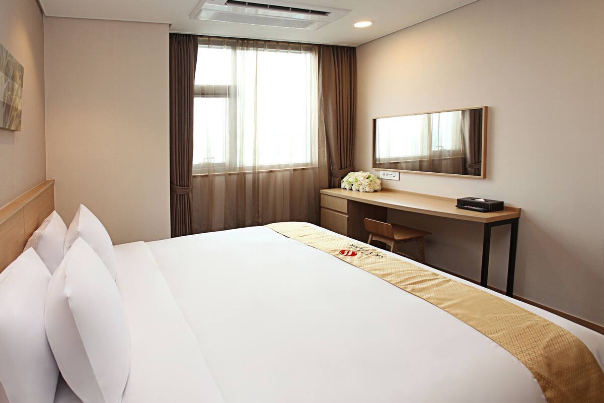 Une chambre d’hôtel comprenant un lit, un bureau, une chaise, une fenêtre avec des rideaux, un miroir et une œuvre d’art accrochée au mur.