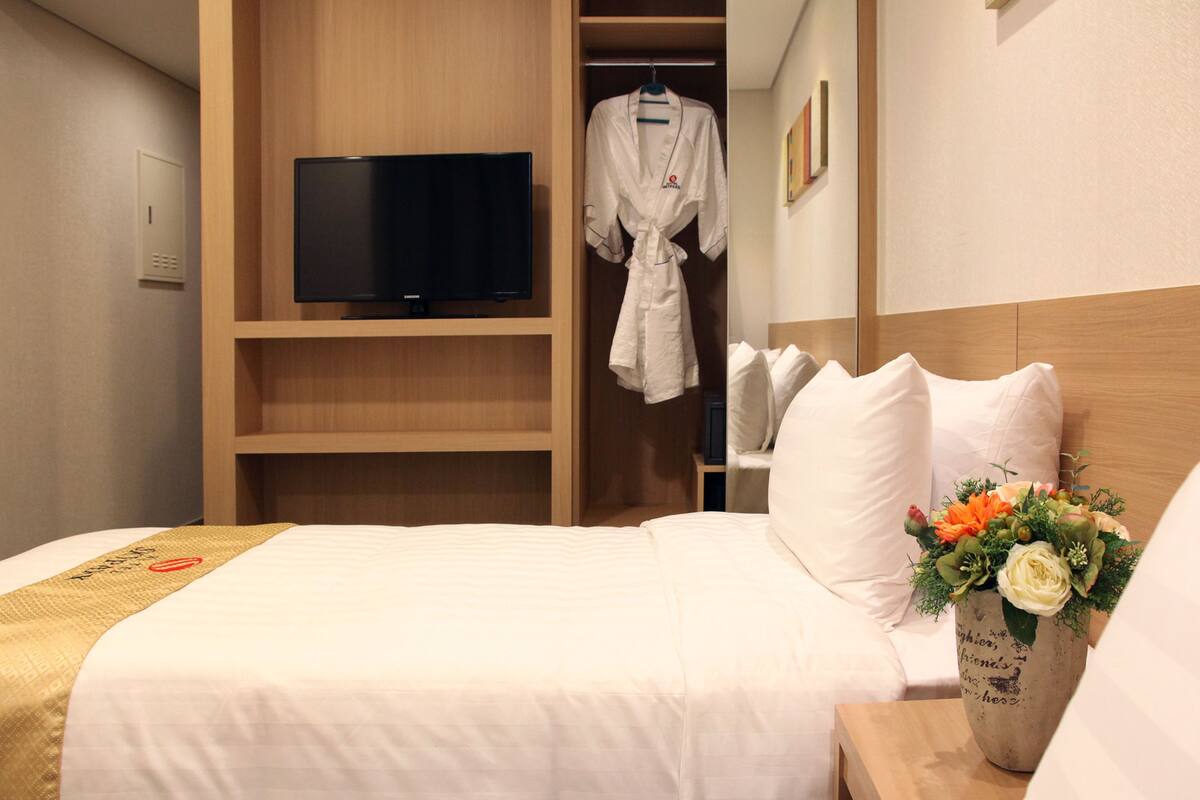 Une chambre d’hôtel avec un lit, une télévision, un peignoir et une table de chevet ornée d’un vase de fleurs.