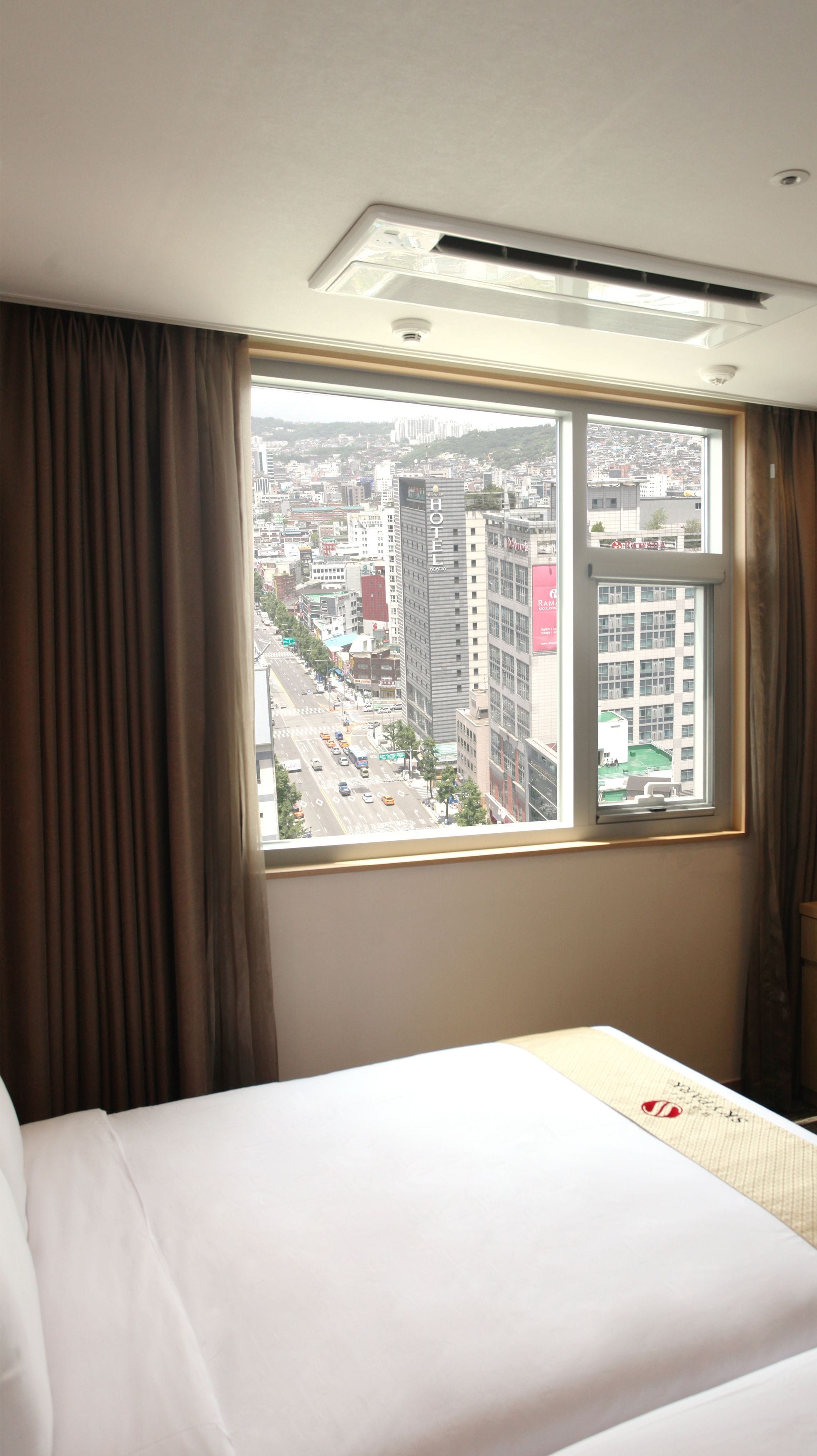 A hotel room with a large window offering a city view, a bed, and a ceiling vent.