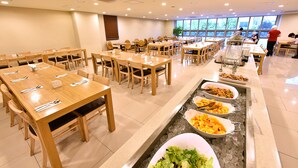 Un comedor amplio con varias mesas y sillas de madera, un bufé con diversas opciones de comida y grandes ventanales que dejan entrar luz natural.