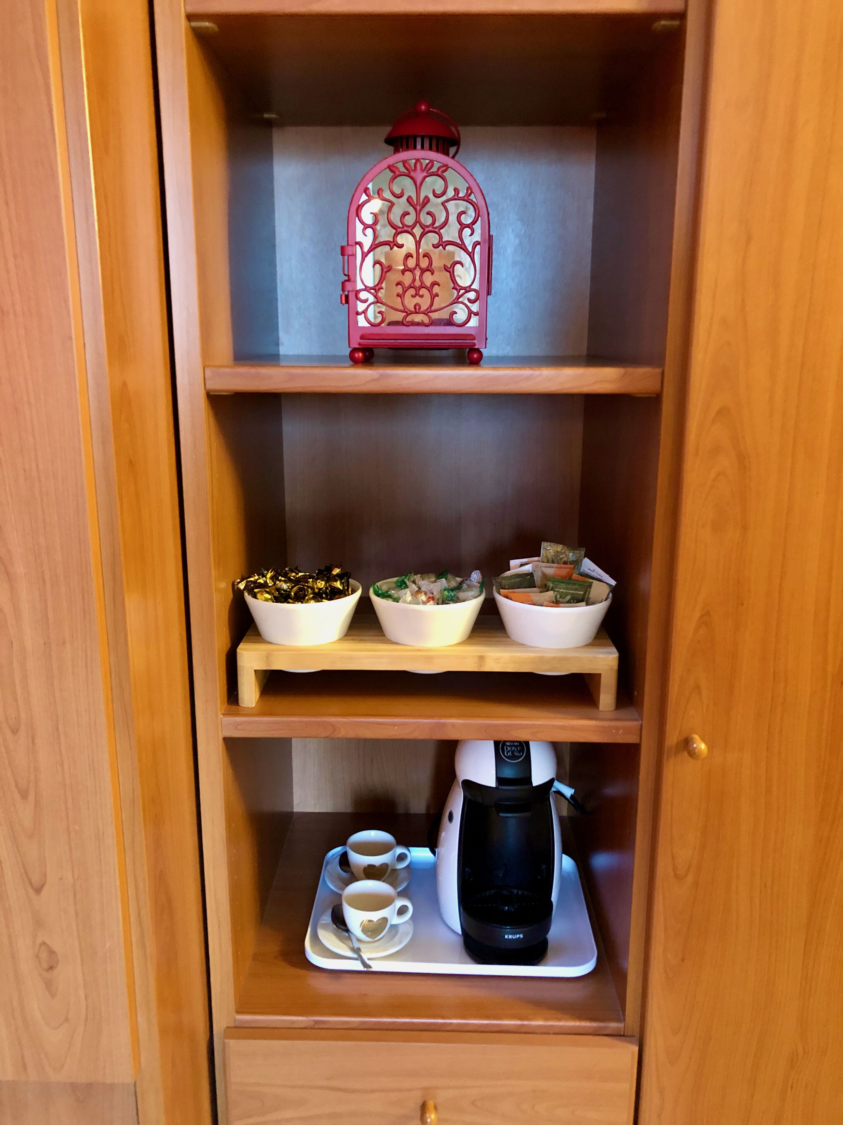 A wooden cabinet with three shelves. The top shelf holds a decorative red lantern. The middle shelf has three bowls with different contents. The bottom shelf has a coffee maker and two cups with heart designs.