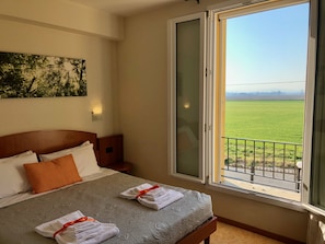 Habitación de hotel con cama, mesita de noche, lámpara de pared y ventana grande con vistas a un campo verde.