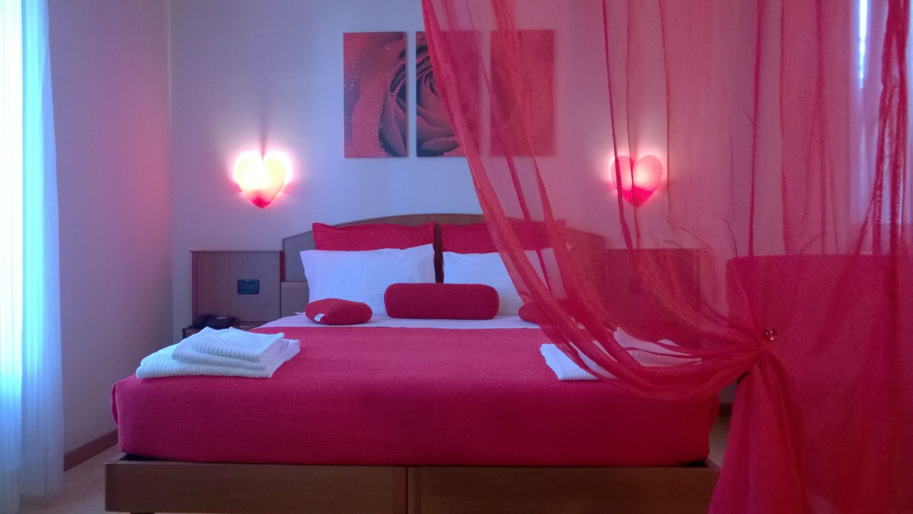 Una cama con ropa de cama roja y almohadas blancas, una mesita de noche y una lámpara fijada a la pared.