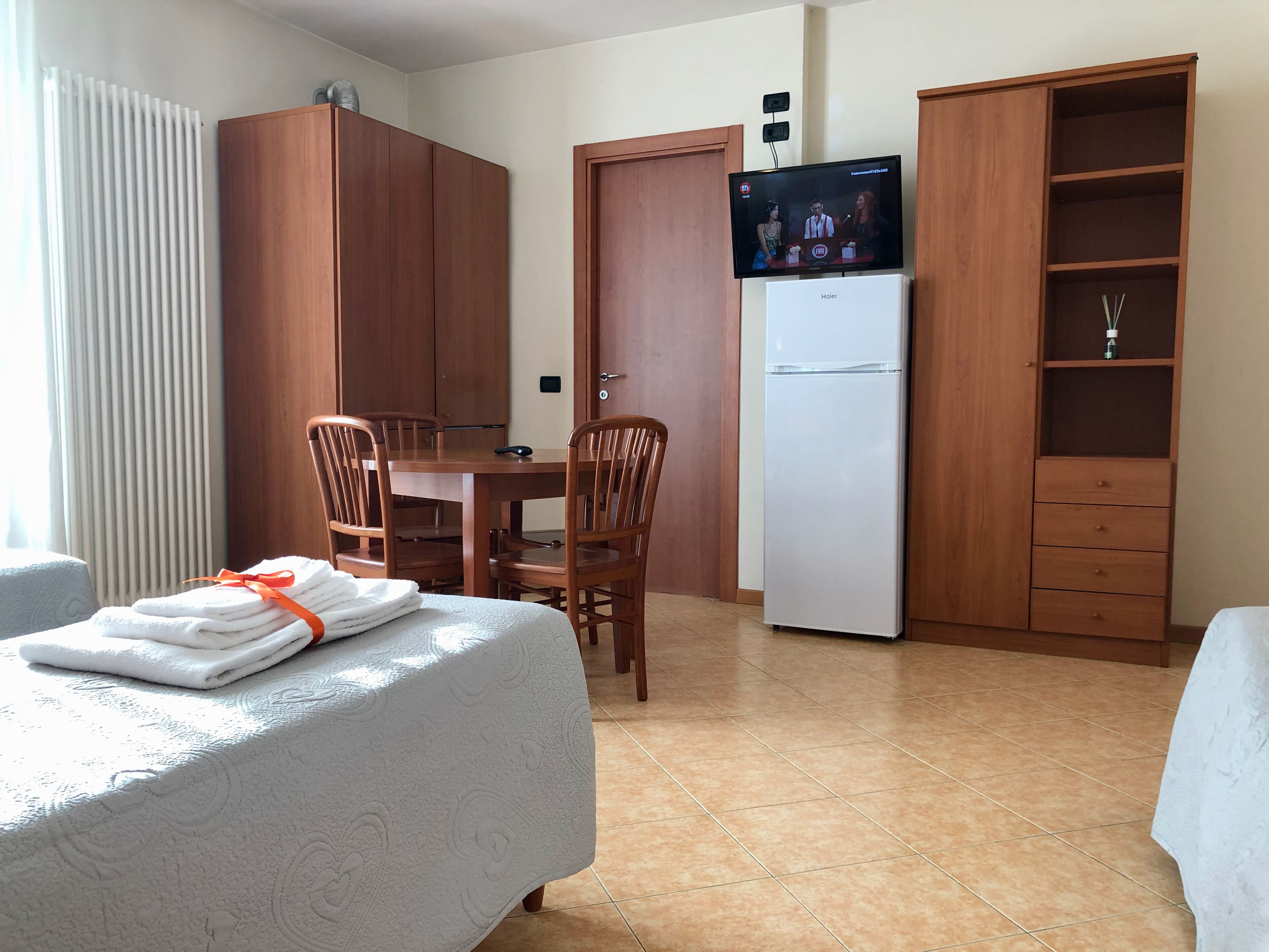 A hotel room with a bed, a dining table with chairs, a white refrigerator, wooden wardrobes, and a television on a stand.