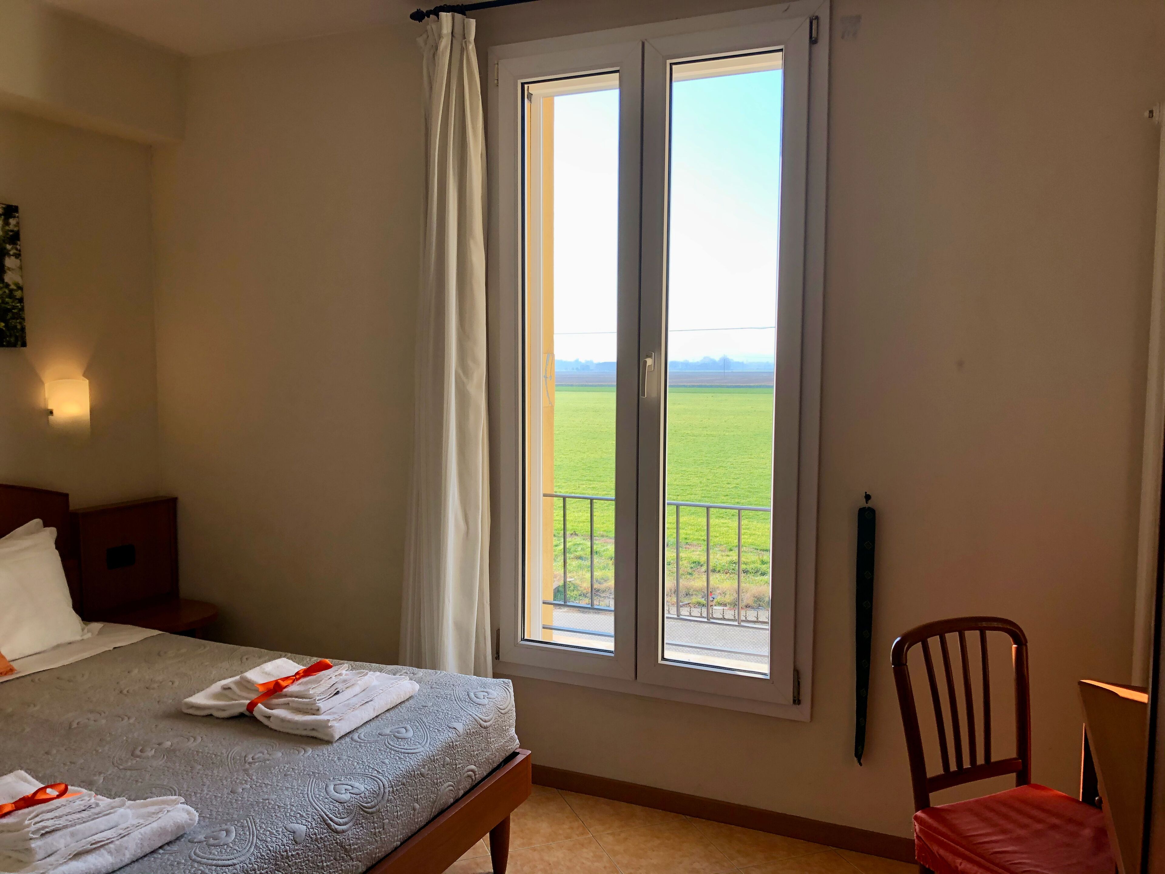 A hotel room with a bed, a chair, a nightstand, and a window with a view of a green field.