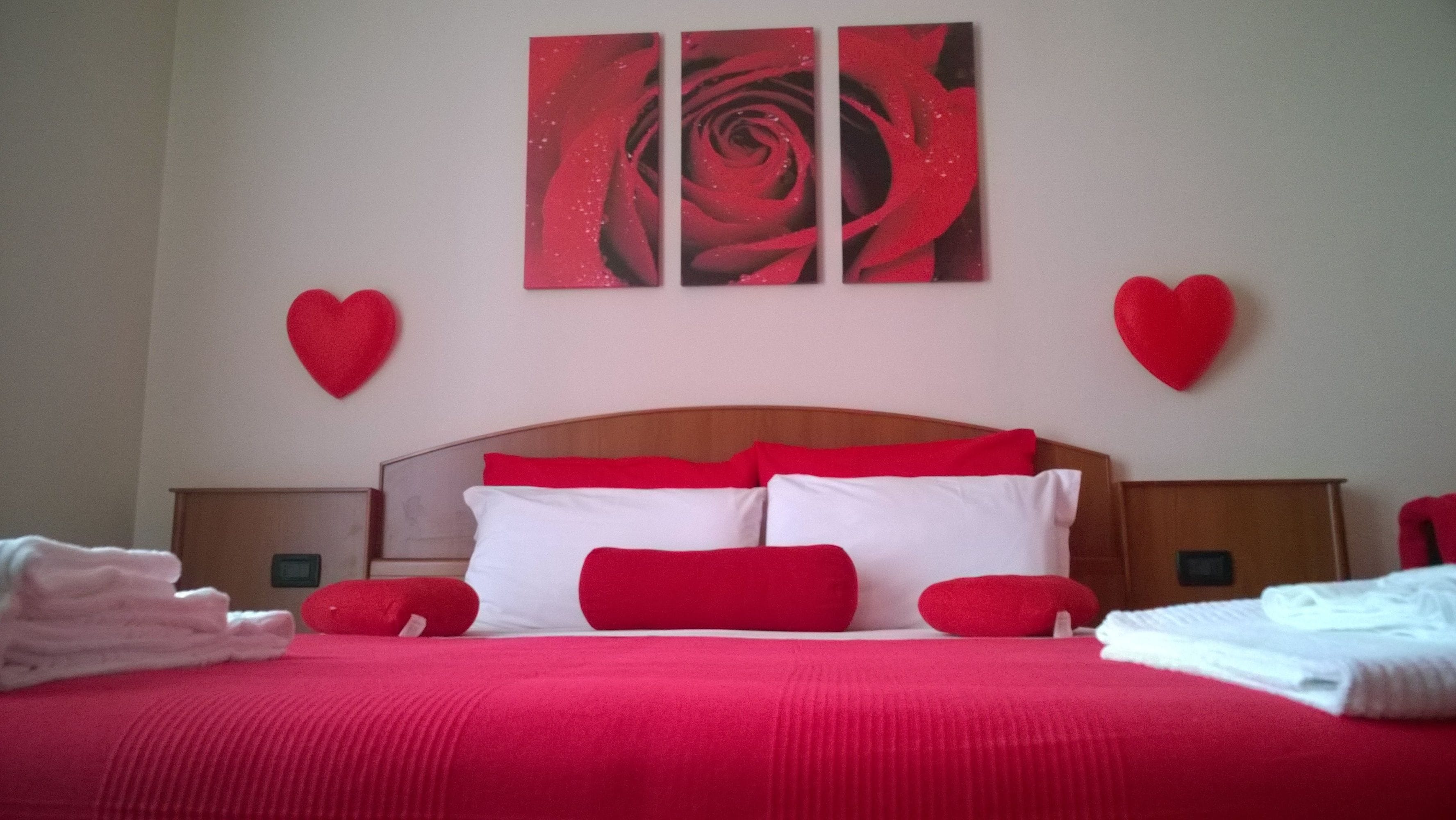 Una habitación de hotel con una cama grande, ropa de cama roja, dos mesitas de noche y una pared con tres cuadros de rosas rojas.
