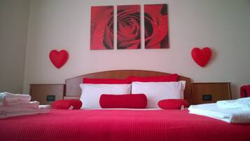 Una habitación de hotel con una cama grande, ropa de cama roja, dos mesitas de noche y una pared con tres cuadros de rosas rojas.