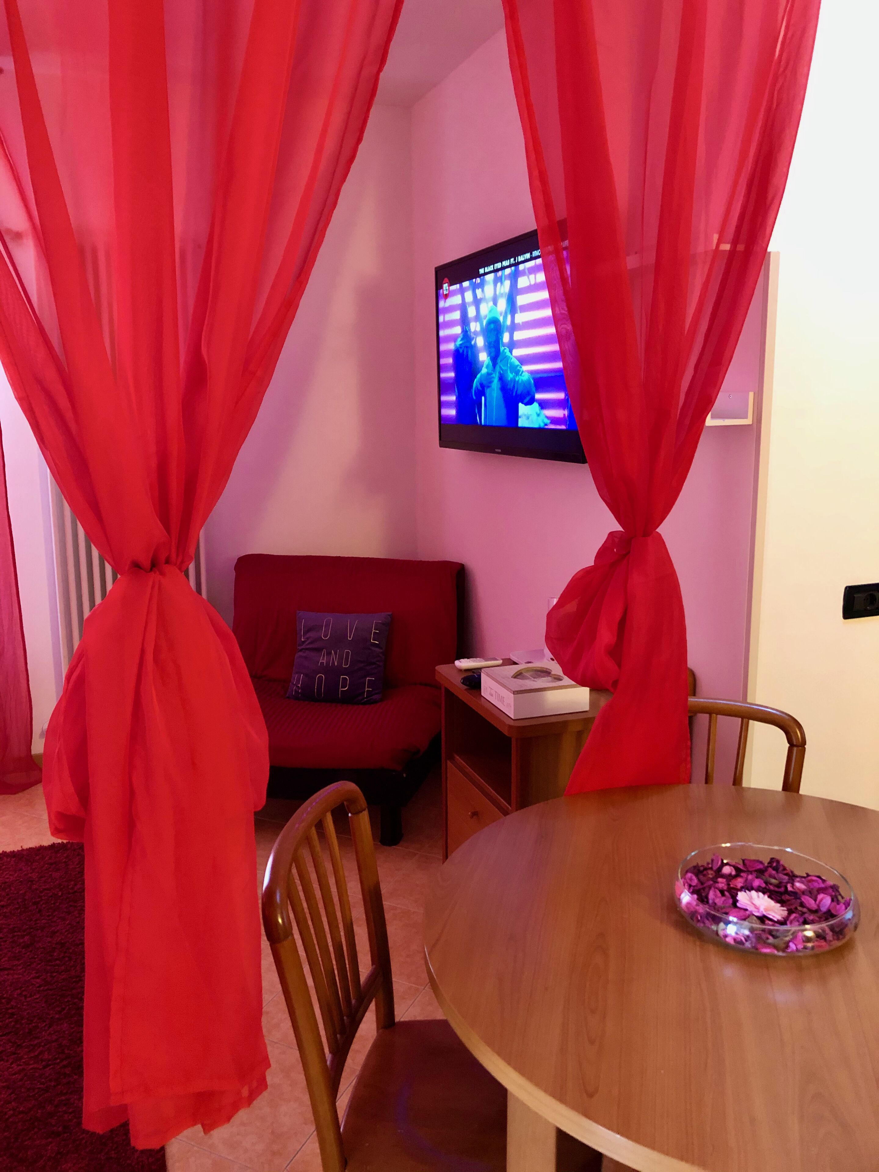 A room with a red sofa, a wooden table with a flower bowl, and a TV on the wall.