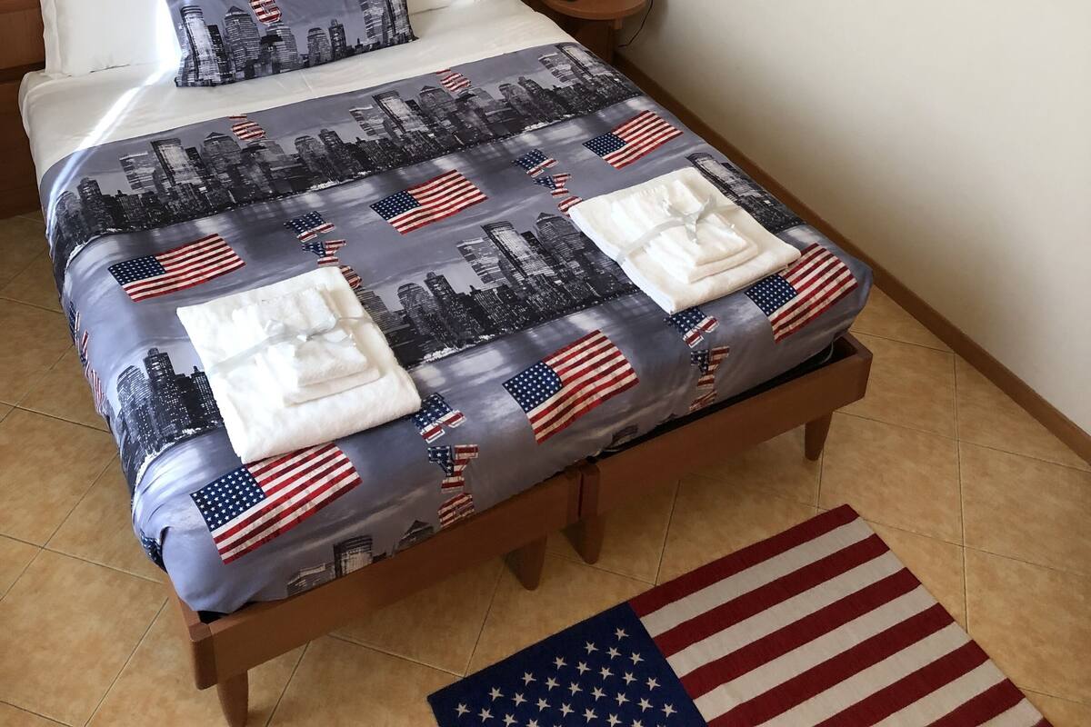 Ein Hotelzimmer mit einem Bett, das ein Stadtbild und ein Muster der amerikanischen Flagge zeigt, einem Nachttisch mit einem Telefon, einer Wand mit verschiedenen Nummernschildern von Bundesstaaten und einem Stadtbild-Kunstwerk über dem Bett.