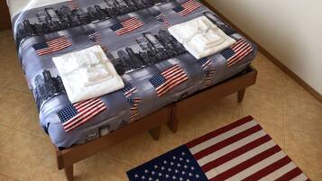 A hotel room with a bed featuring a cityscape and American flag pattern, a nightstand with a phone, a wall with various state license plates, and a cityscape artwork above the bed.