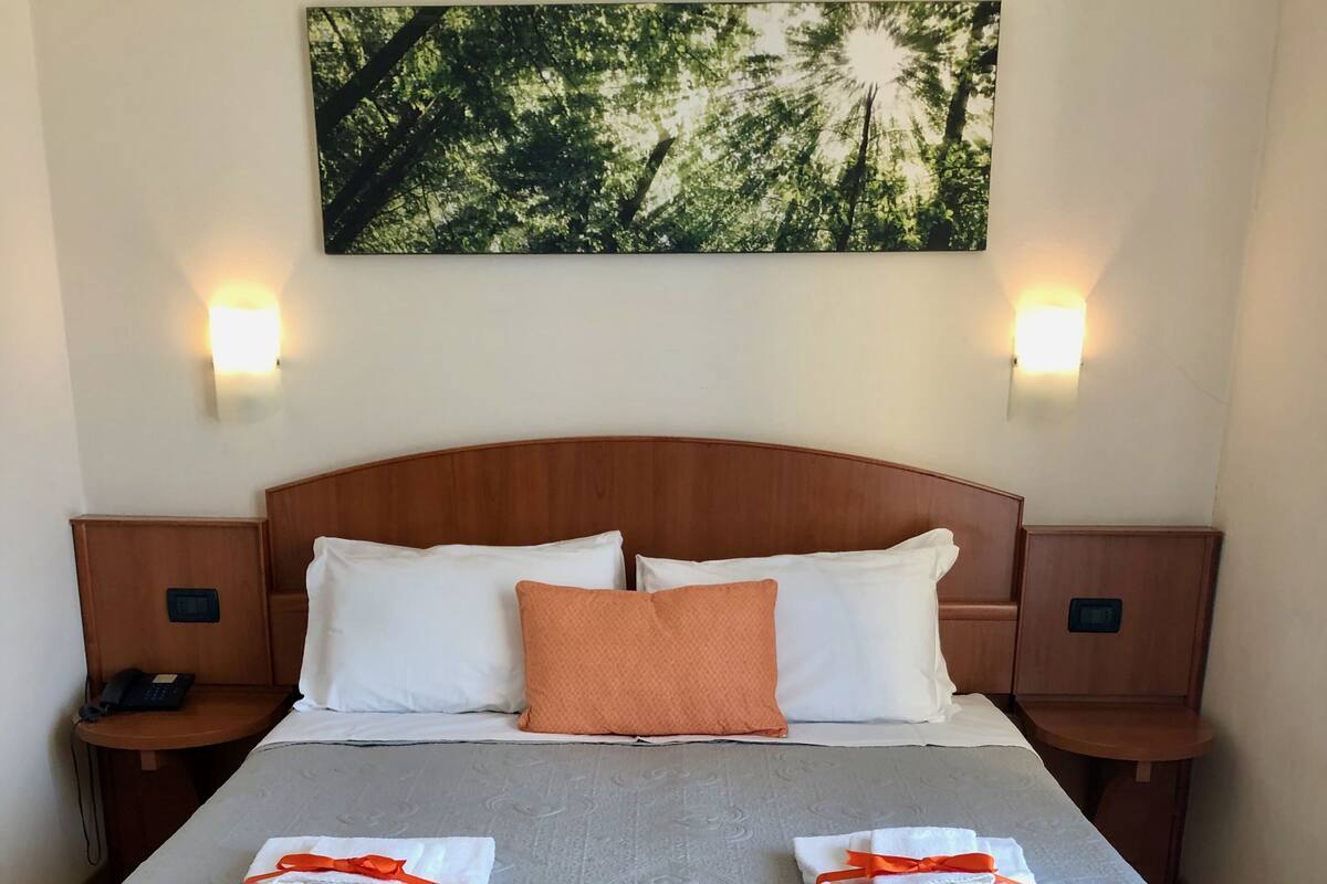Ein ordentlich bezogenes Bett mit einer grauen Decke, zwei weißen Kissen und einem orangefarbenen Zierkissen. Es gibt zwei Nachttische mit Lampen, ein an der Wand montiertes Telefon und ein gerahmtes Bild über dem Bett.