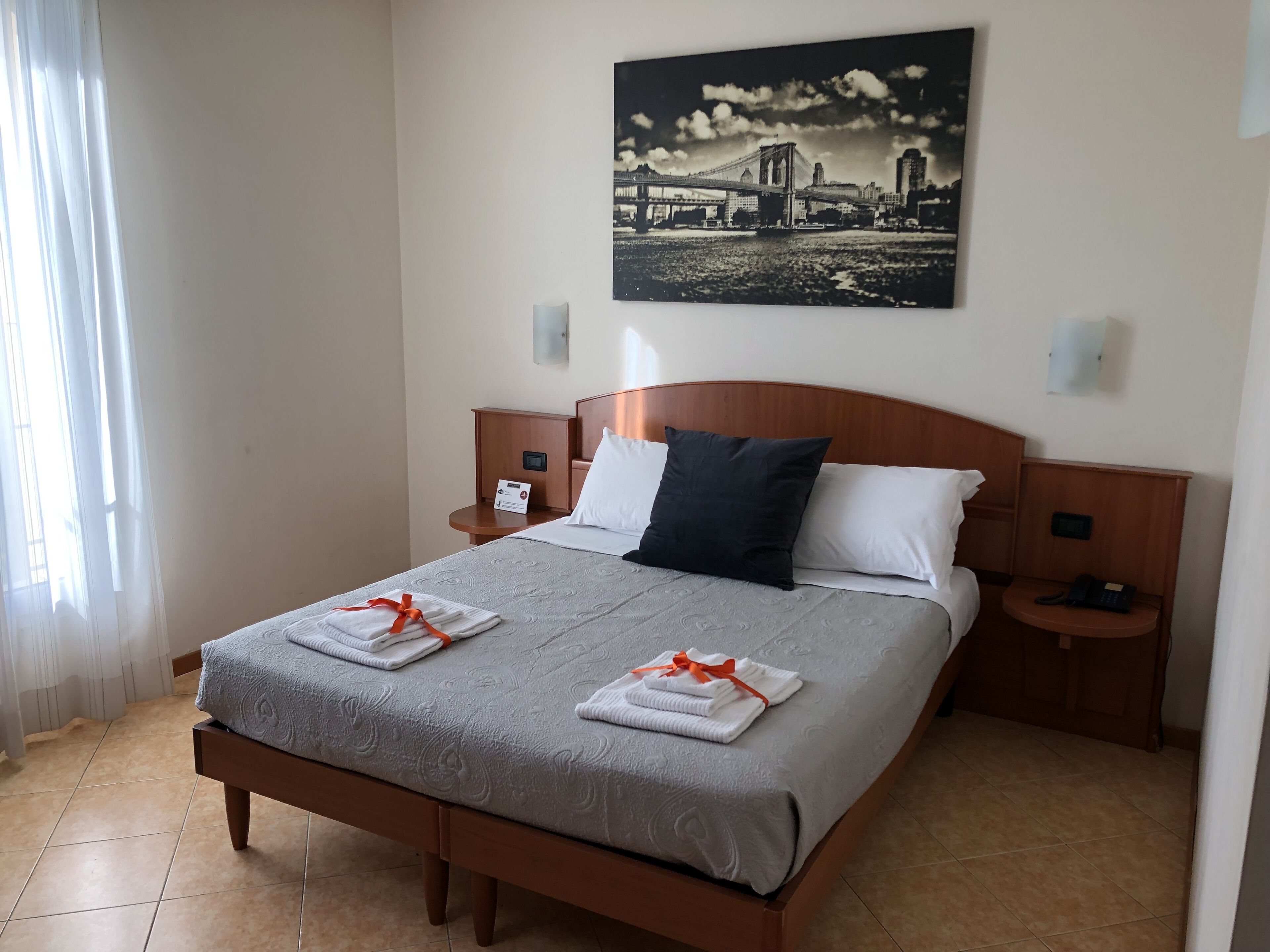 A neatly made bed with a grey bedspread and white pillows, two bedside tables with lamps, a framed picture of a bridge on the wall, and a window with sheer curtains.