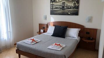 A neatly made bed with a grey bedspread and white pillows, two bedside tables with lamps, a framed picture of a bridge on the wall, and a window with sheer curtains.