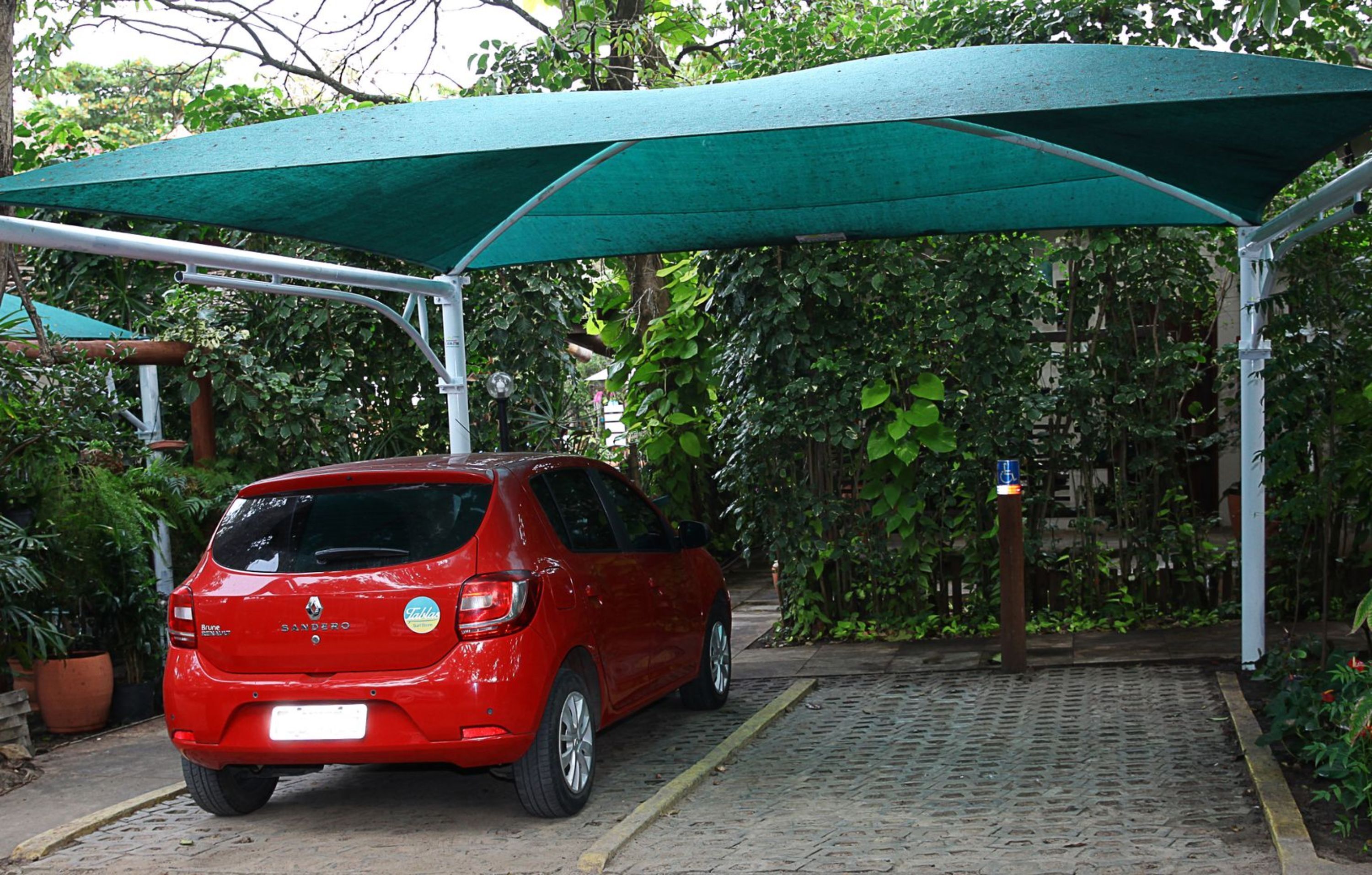 A red car parked under a green canopy in a covered parking area with a cobblestone path.