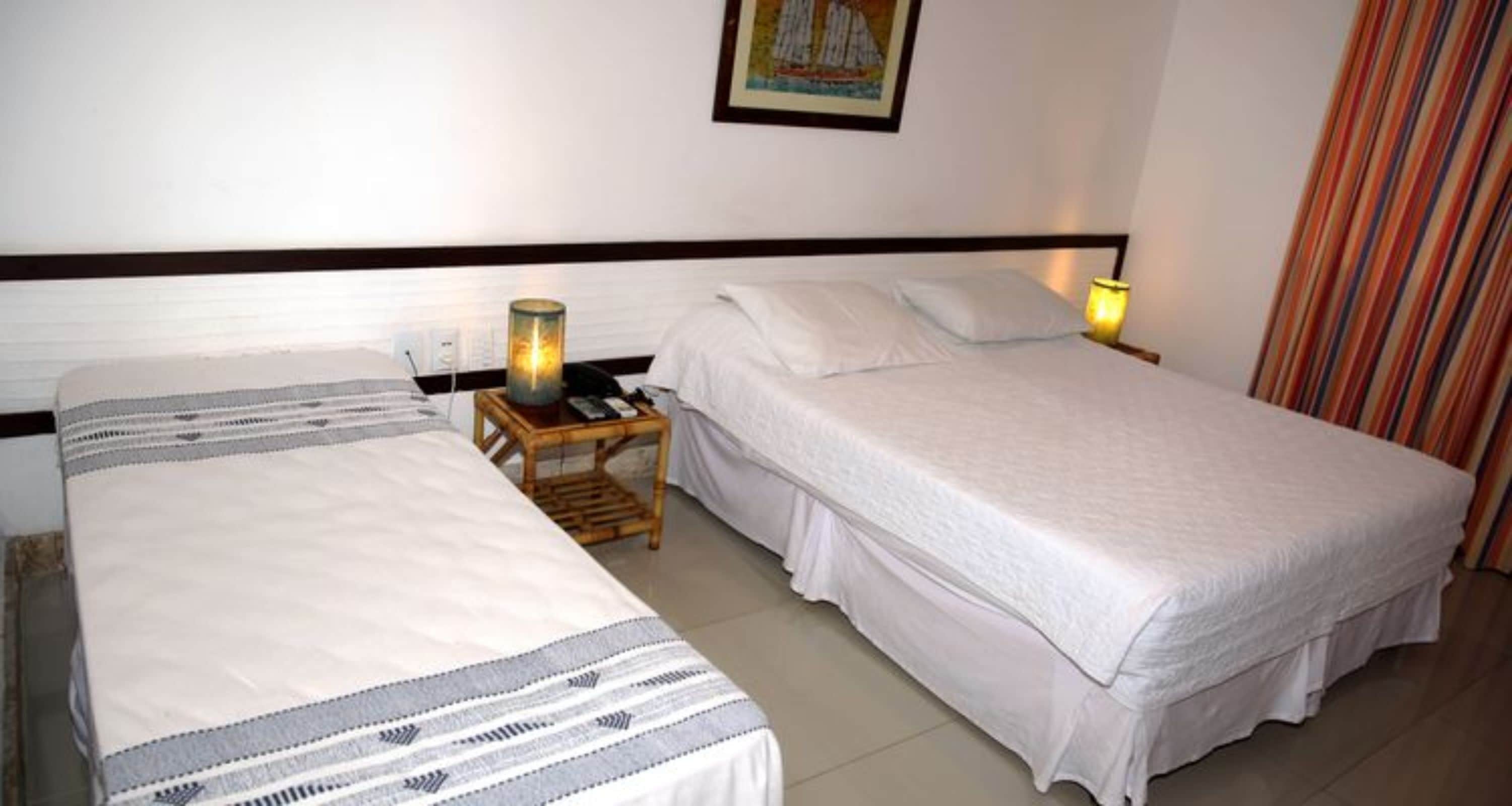 Two single beds with white linens, a wooden nightstand with a lamp, and a striped curtain.