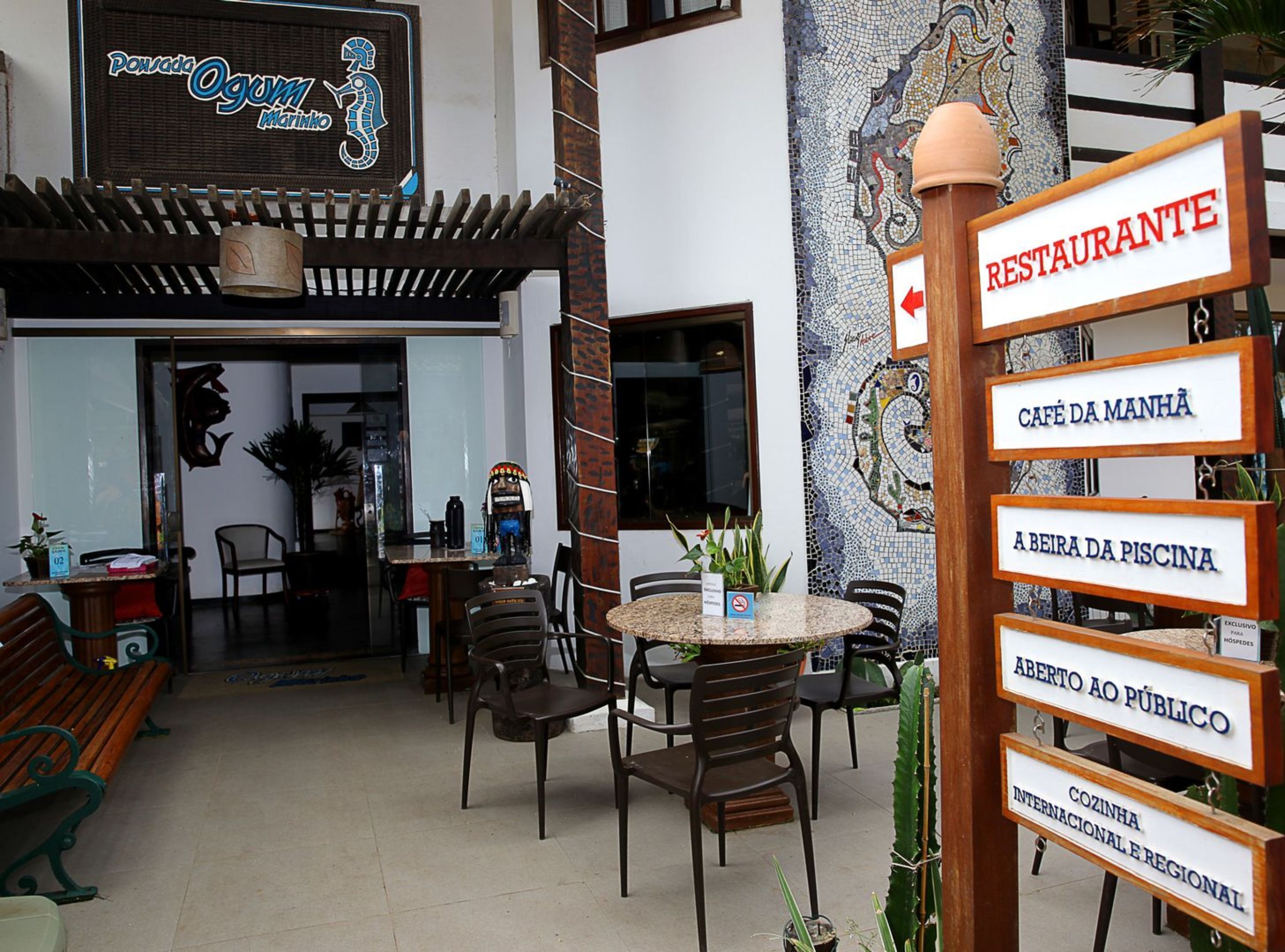 An outdoor seating area with tables and chairs, a mosaic wall, and a sign indicating a restaurant.