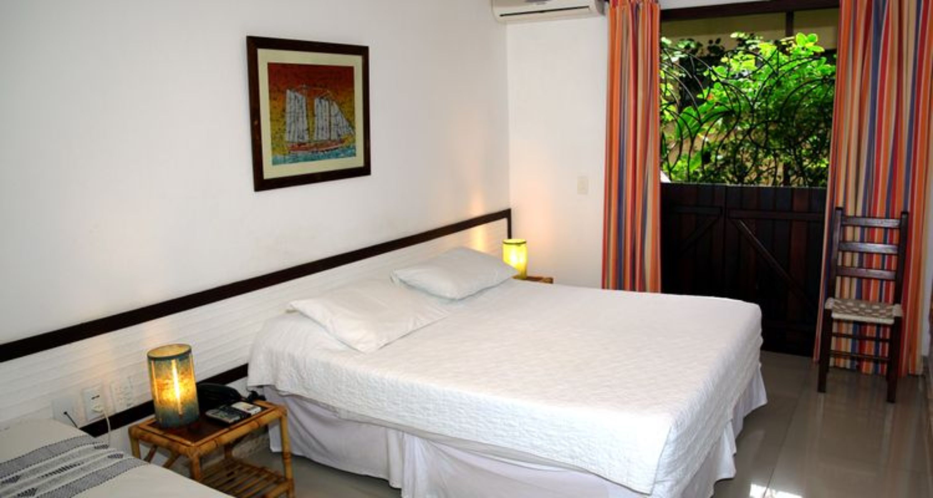 Een hotelkamer met een bed, een nachtkastje met lamp, een ingelijst schilderij aan de muur en uitzicht op groen door een deur.