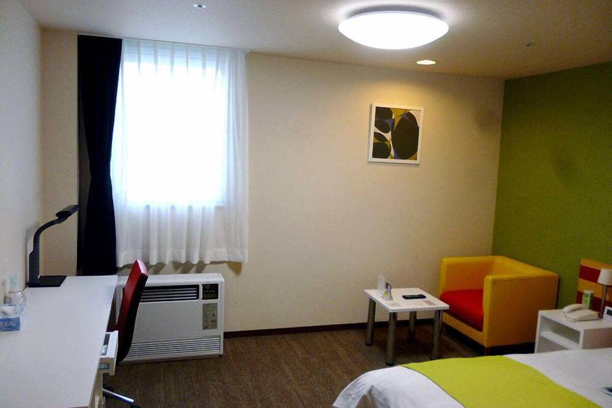 Ein Hotelzimmer mit Bett, Schreibtisch, Stuhl, einem kleinen Tisch, einer Klimaanlage und einem Fenster mit Vorhängen.