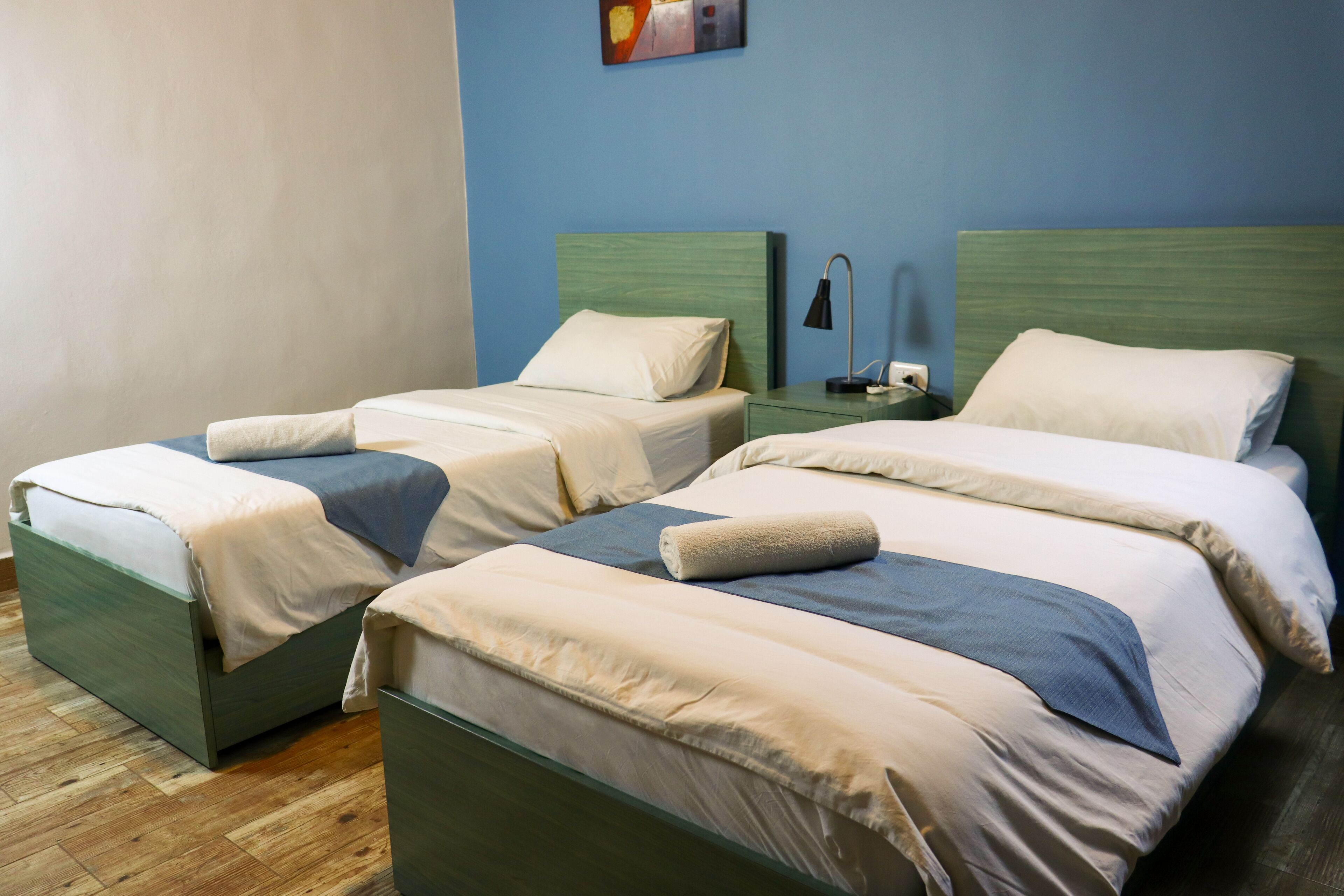 Una habitación de hotel con dos camas individuales, cada una con cabecero de madera, una mesita de noche con lámpara y pared azul.