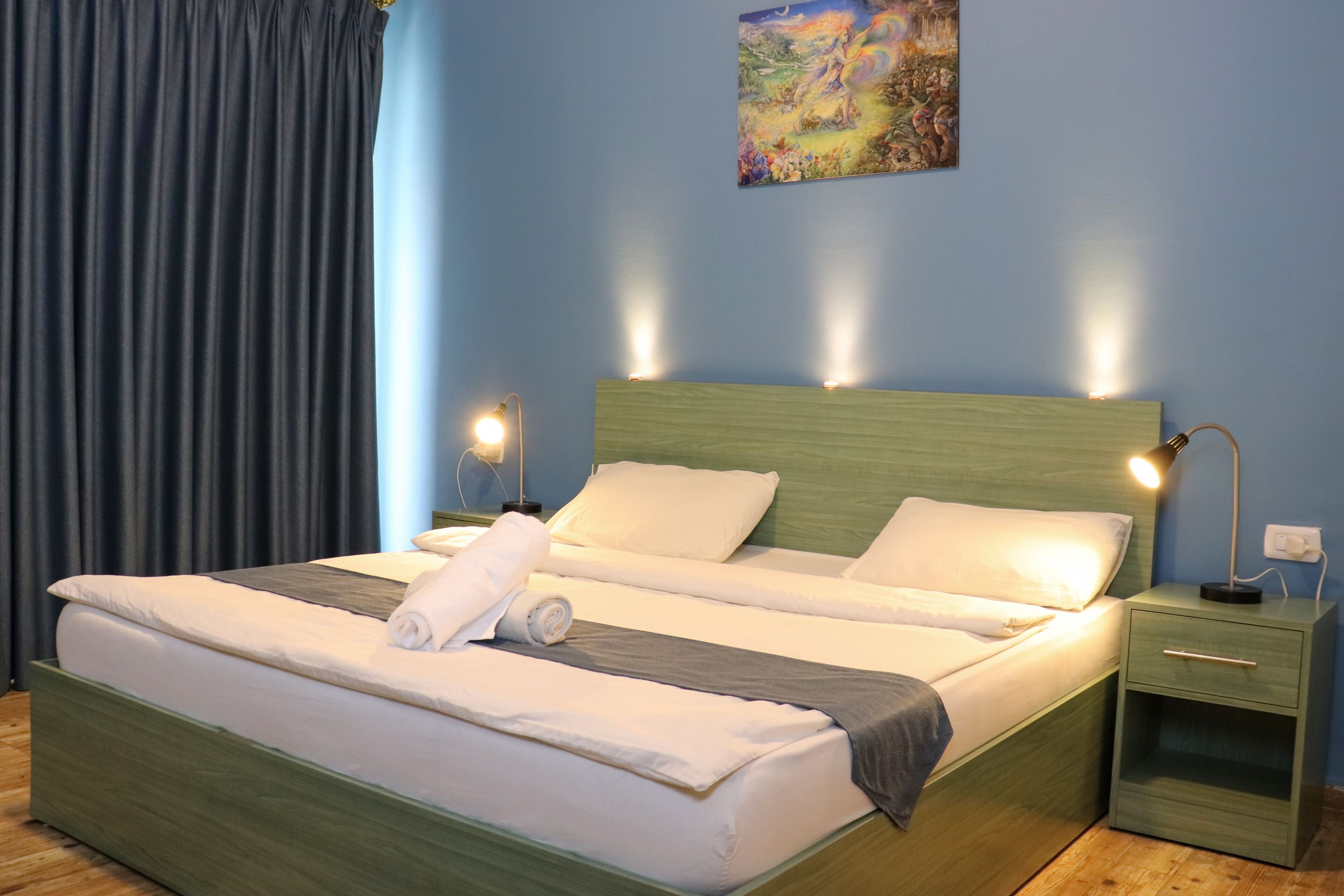 A neatly made bed with white linens and a rolled towel, a green nightstand with a lamp, and a painting on the wall.