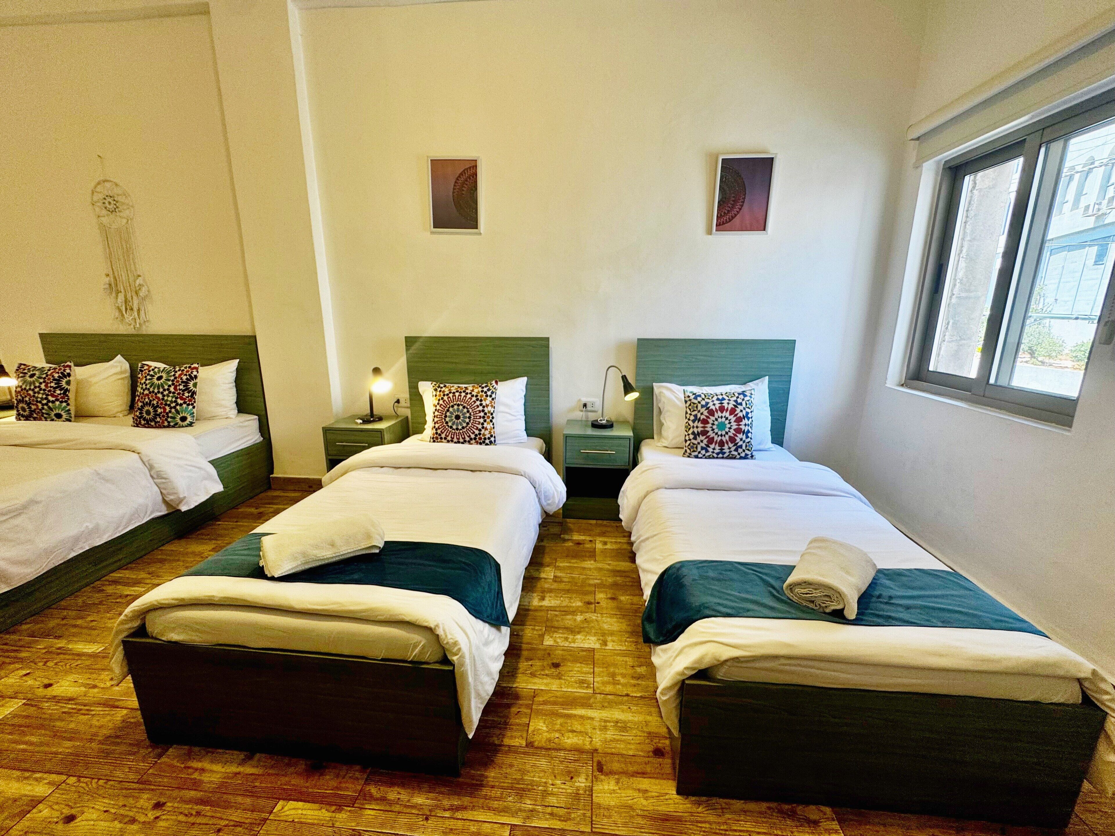 A hotel room with three single beds, wooden flooring, and a window providing natural light.
