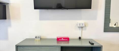 A hotel room with a flat-screen TV mounted on the wall, a small desk with drawers, a trash bin, and a minibar.