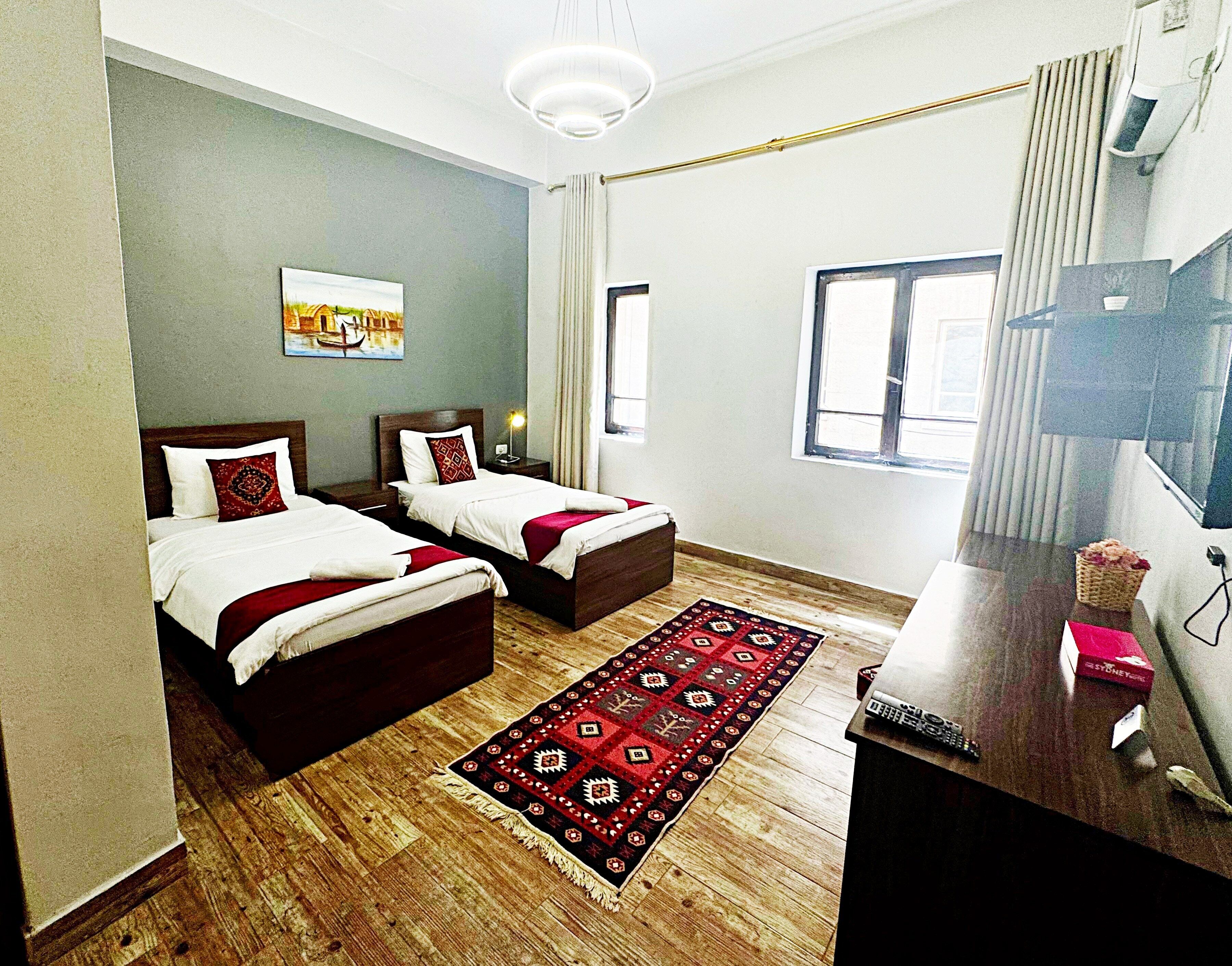 A hotel room with two beds, a wooden floor, a red and black patterned rug, a desk with a computer and a box, and a window with curtains.