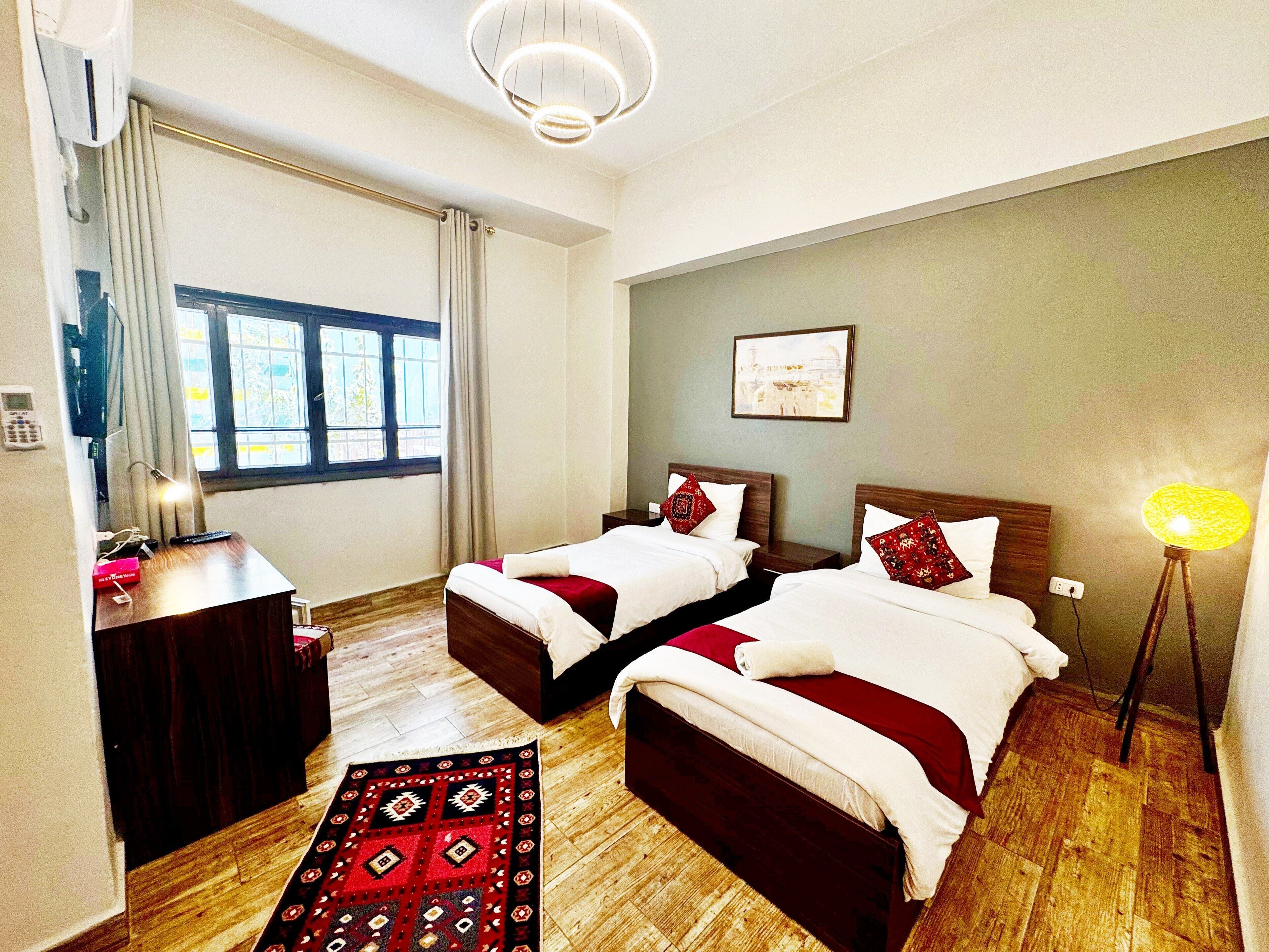 A hotel room with two beds, a wooden floor, a red and black patterned rug, a desk with a lamp, and a window with curtains.