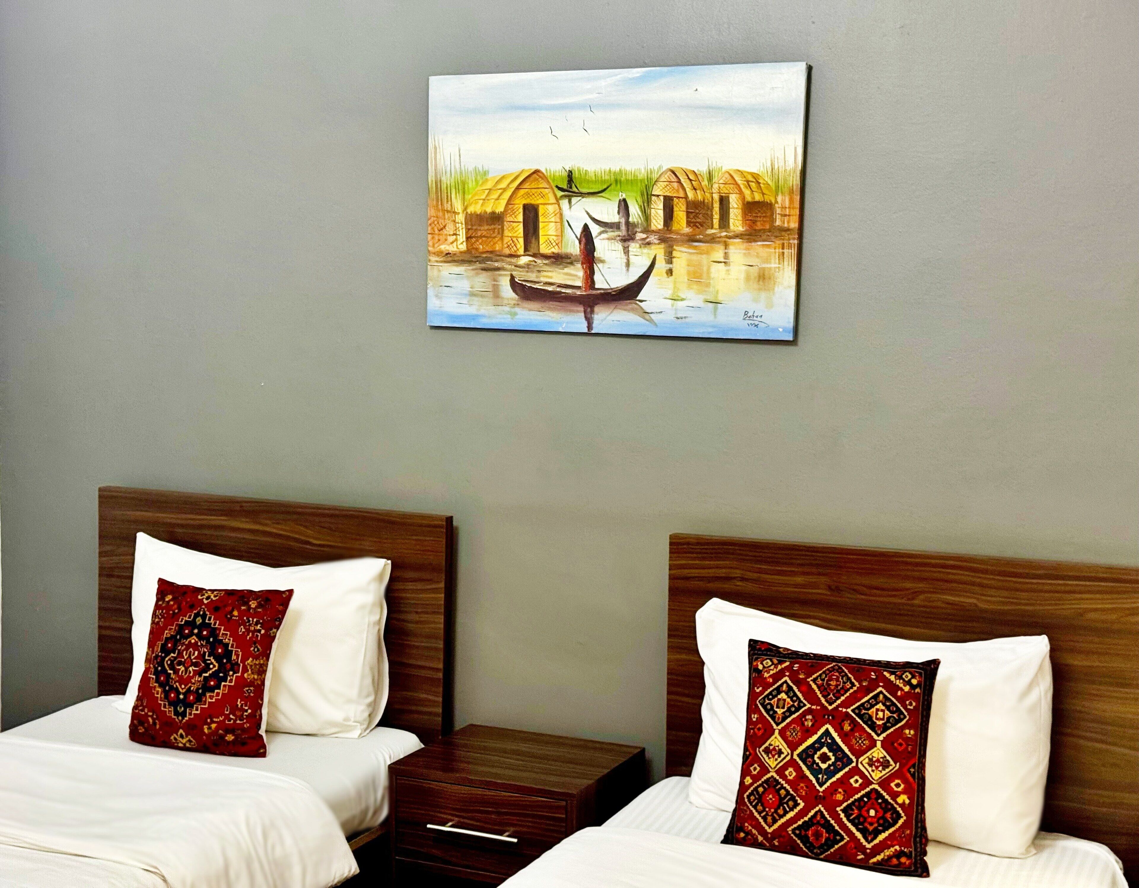 A hotel room with two beds, each with a wooden headboard and white bedding. A painting of a traditional scene hangs on the wall above the beds.