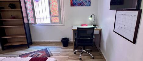A bedroom with a bed, a desk, a chair, a window with patterned glass, an air conditioner, and a wall-mounted TV.