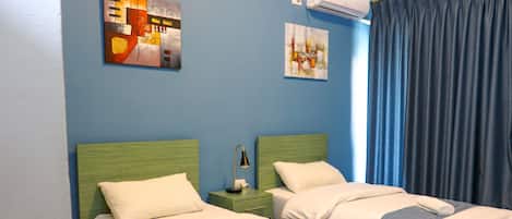 A hotel room with two beds, each with a green headboard, white bedding, and a rolled towel. There are two paintings on the wall, a nightstand with a lamp, and a window with blue curtains.