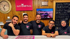 Un grupo de personas en la recepción de un hotel en Sydney, con un reloj, un televisor y una pizarra con información del hotel.